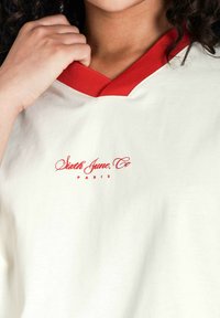 Camisa color crema con un cuello V rojo. Presenta texto rojo que dice "Sixth June Co Paris" en la parte frontal. Tejido suave, diseño casual.