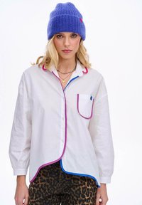 Biała koszula z różowym i niebieskim wykończeniem, długie rękawy, detal kieszeni, oraz spodnie w panterkę. Modelka nosi fioletową dzianinową czapkę i warstwowe naszyjniki.