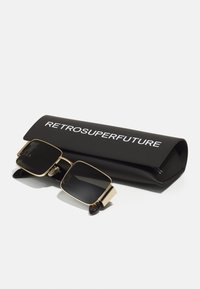 Lunettes de soleil avec des verres noirs et une monture métallique dorée, reposant sur un étui rectangulaire noir avec "RETROSUPERFUTURE" imprimé en blanc.