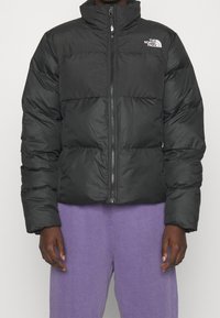 Osoba ubrana w czarną kurtkę puchową z logo The North Face oraz fioletowe spodnie, stojąca z rękami luźno opuszczonymi wzdłuż ciała.