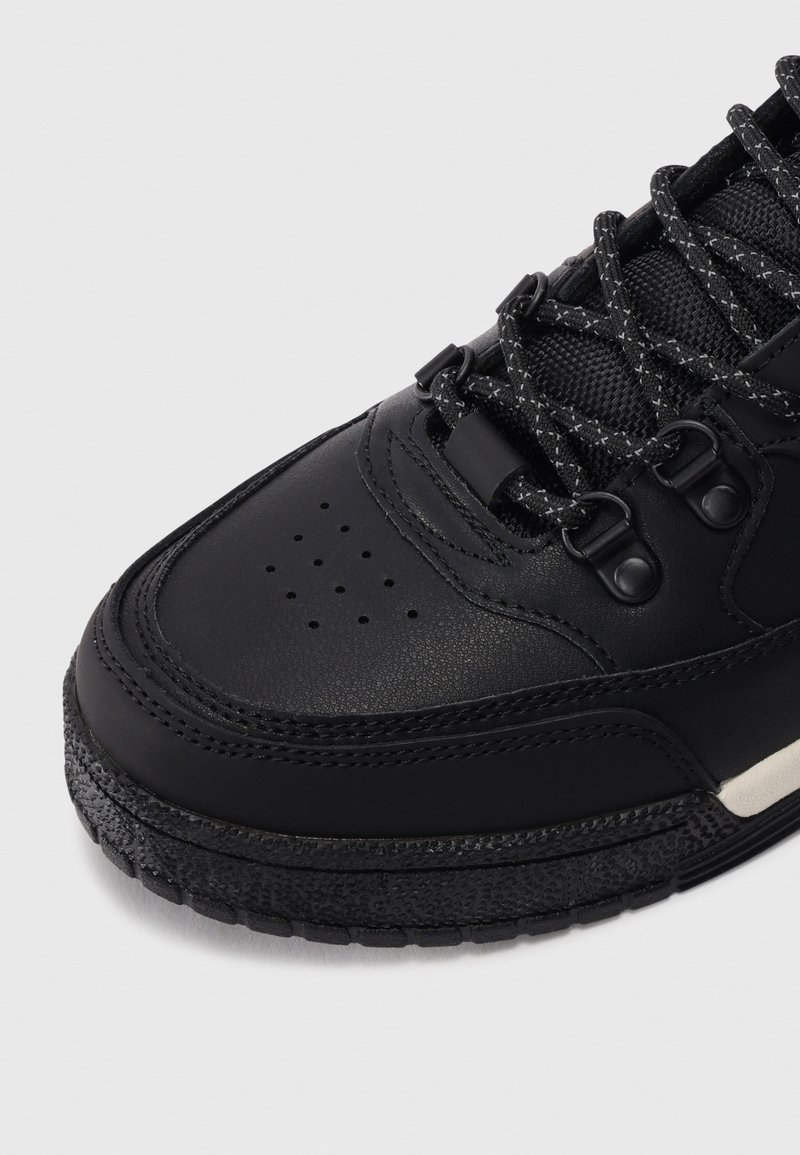 Schwarzer Leder-Sneaker mit strukturiertem Sohlenbereich, schwarz-graue gemusterte Schnürsenkel und Metallösen auf einem weißen Hintergrund.