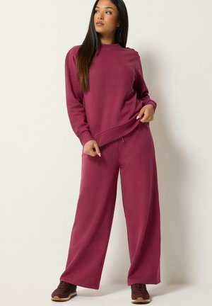 Kvinde iført matchende løstsiddende vinrød sweatshirt og vidde bukser med brune sneakers, stående mod en ensfarvet lys baggrund.
