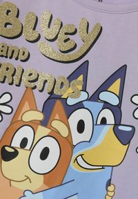 Fioletowa koszulka z błyszczącym złotym napisem "Bluey i Przyjaciele". Dwa bajkowe psy, jeden pomarańczowy i jeden niebieski, z dużymi oczami i prostymi rysami twarzy.