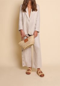 Kvinna i beige linnedräkt med vida byxor med knappar, hållande en vävd stroppväska och iförd beiga sandaletter med spänne.