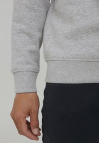 Grijze katoenen sweatshirt met ribgebreide manchetten en zoom, met een gladde textuur. Hand zichtbaar, rustend aan de onderkant van het kledingstuk.