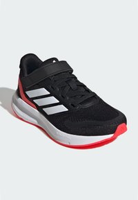 Chaussure de sport noire avec une tige en mesh, des rayures blanches et une semelle rouge. Dispose d'une sangle et d'un col rembourré pour le confort et le soutien.