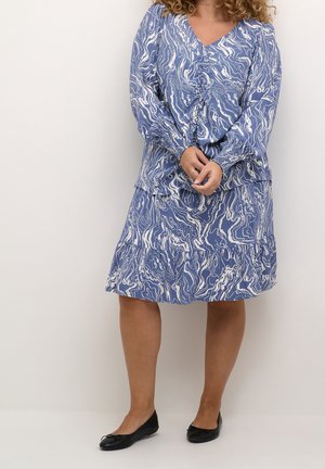 Vrouw die een blauw-witte marmeren blouse met lange mouwen en een bijpassende rok draagt, staand met gevouwen handen en zwarte ballerina's.