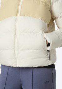 Person som bär en färgblockerad beige och vit vadderad jacka med händerna i fickorna, i kombination med blå North Face-byxor.