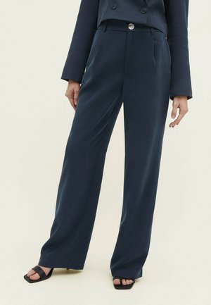 Vrouw draagt donkerblauwe, wijd uitlopende broek met hoge taille en een dubbelgeknoopt donkerblauw blazerjasje, gecombineerd met zwarte open sandaalhakken.