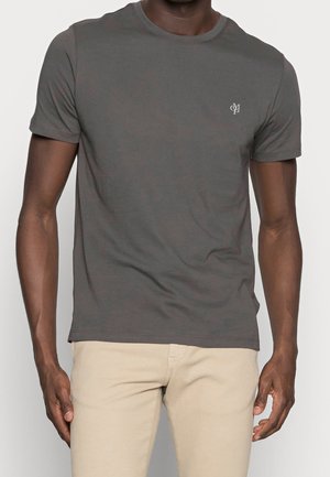 Man draagt een aansluitend donkergrijs T-shirt met korte mouwen en een klein wit logo op de borst, gecombineerd met beige broeken tegen een effen achtergrond.