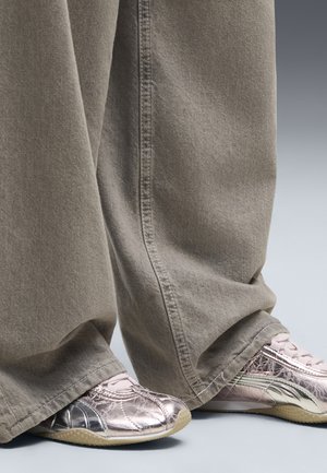 Pantaloni beige larghi alla lunghezza della gamba che coprono parzialmente sneakers rosa metallizzato con suole beige su un pavimento grigio chiaro.