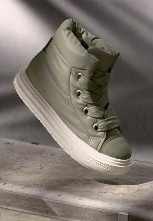 Wysokie, wyściełane trampki w stonowanym zielonym kolorze z grubymi sznurowadłami i białą gumową podeszwą unoszące się nad betonową powierzchnią.