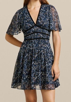 Donna che indossa un vestito corto blu scuro a fiori con maniche leggere e trasparenti e una scollatura profonda a V, in piedi con una mano sul fianco.