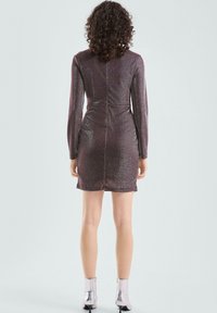 Kobieta z kręconymi włosami, ubrana w błyszczącą purpurową mini sukienkę z długim rękawem i srebrne botki za kostkę, stojąca plecami na jednolitym tle.