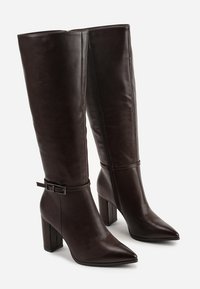 Bottes en cuir marron foncé arrivant jusqu'au genou, avec un bout pointu, un talon épais et un accent de boucle au niveau de la cheville. Texture lisse sur l'ensemble.