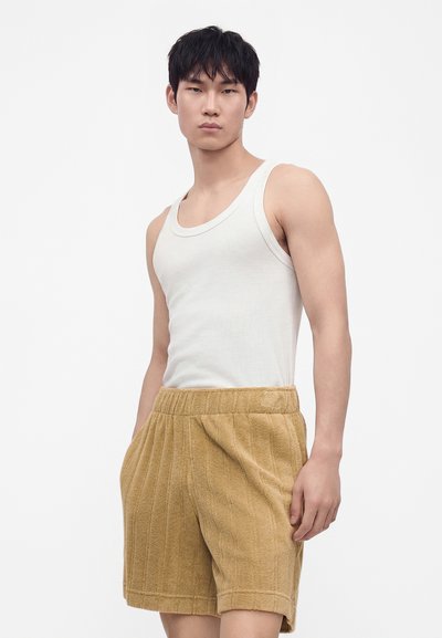Jeune homme portant un débardeur blanc et un short marron clair texturé, debout les mains dans les poches devant un fond uni.