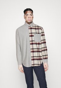 Hemd mit gestreifter linker Seite und kariertes Design auf der rechten Seite in Beige, Rot und Schwarz. Auffälliges Taschendetail im karierten Bereich.