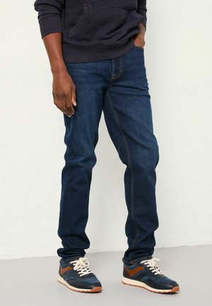 Person trägt dunkelblaue Jeans, marineblaue und braune Sneakers und einen dunklen Hoodie und steht mit einer Hand in der Tasche an einer weißen Wand.
