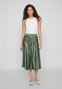 Gonna plissettata verde in satin con una cintura elastica ampia, abbinata a un top senza maniche bianco e scarpe slip-on di colore chiaro.