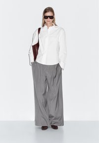 Donna che indossa una camicia bianca oversize, pantaloni larghi grigi, stivali marrone scuro, grandi occhiali da sole e porta una borsa a tracolla marrone scuro.