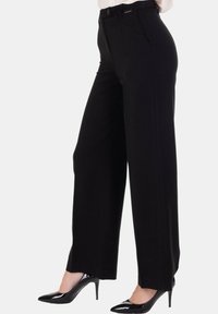 Pantaloni neri a gamba larga indossati con scarpe col tacco alto in vernice nera, mostrati dalla vita ai piedi su uno sfondo semplice.