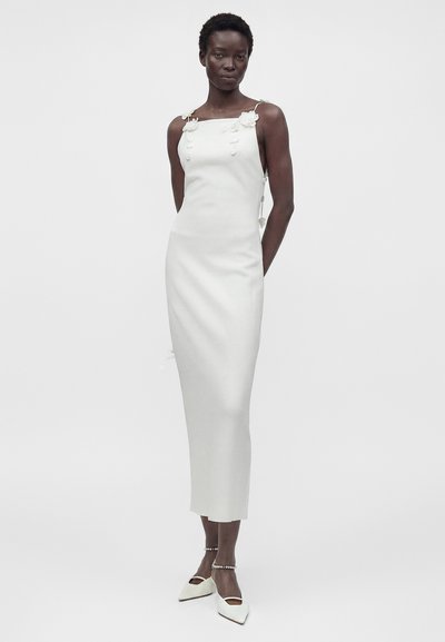 Γυναίκα φοράει άσπρο μίντι φόρεμα χωρίς μανίκια με λουλουδάτες τιράντες και άσπρα μυτερά παπούτσια με λουράκια στον αστράγαλο, στέκεται μπροστά σε απλό φόντο.