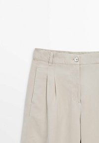 Beige pants featuring a smooth texture, pleated front design, button closure, and belt loops. Tailored cut for a structured look.