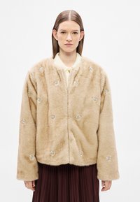 Casaco de pele sintética bege com textura macia, decorado com apliques de flores prateadas e corte descontraído. Gola baixa e mangas compridas.