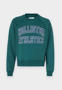 Felpa verde acqua con polsini e orlo a coste, con grande scritta "HOLLISTER ATHLETICS" bianca e blu scuro sulla parte anteriore. Realizzata in tessuto morbido.