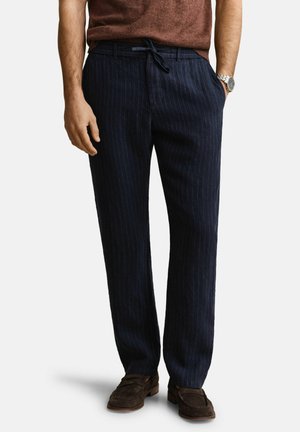 Mann trägt marineblaue Nadelstreifenhose mit Kordelzugbund, braune Anzugschuhe und einen braunen Pullover, Hände teilweise in den Taschen.