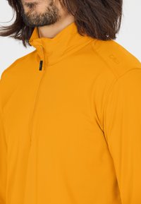Letvægtig, kvart-zip trøje i lys gul materiale med en glat tekstur. Har en fold-down krave og subtil branding på skulderen.