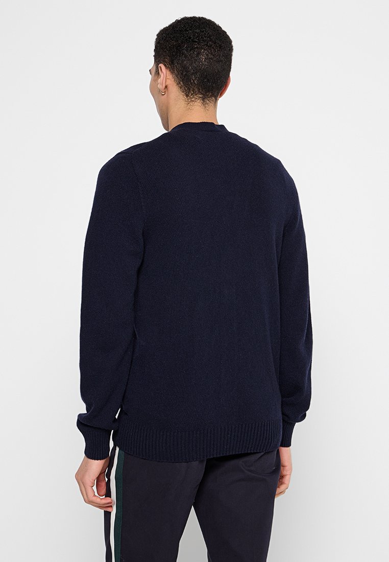 Uomo con capelli corti e ricci, che indossa un maglione blu navy e pantaloni scuri con una striscia laterale, in piedi di spalle su uno sfondo bianco.