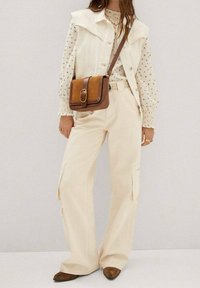 Beige weite Cargo-Hosen, cremefarbene ärmellose Jacke mit Kragen, weiße Bluse mit blauen Mustern, braune Umhängetasche und braune Schuhe.