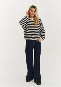 Dziergany sweter w paski w beżowym i granatowym kolorze, o luźnym kroju i szerokich rękawach, zestawiony z ciemnoniebieskimi dzwonami i czarnymi butami.