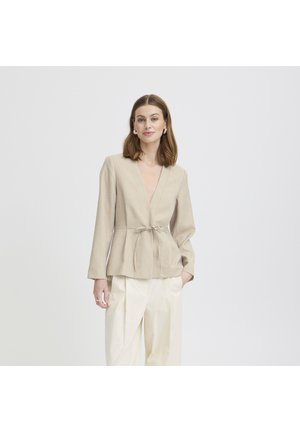 Kvinde iført beige slå-om-langærmet top med bindebånd i taljen og løse hvide bukser, stående med den ene hånd i lommen mod en hvid baggrund.