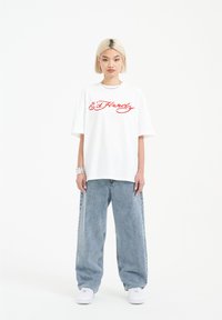 Bílé oversized tričko s červeně vyšitým logem "Ed Hardy", v kombinaci s světle modrými širokými džíny a bílými teniskami. Jednoduchý design.