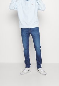 Sudadera con capucha azul claro con un logo rojo, combinada con jeans ajustados azules y zapatillas blancas con detalles en negro. Textura suave, diseño casual.