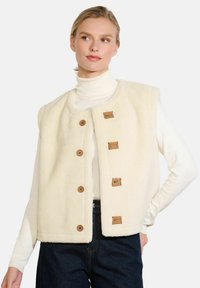 Blonda sieviete, tērpta krēmkrāsā fliša vestē ar koka pogām, virs balta turtlenecka un tumšām džinsām, pozē ar vienu roku uz gūžas.