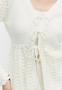 Biała dziergana kamizelka o prążkowanej fakturze i koronkowych detalach. Posiada dekolt w kształcie litery V i wiązania z przodu. Długie, wzorzyste rękawy.