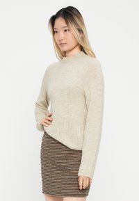 Vaalean beige neulepusero korkealla kauluksella ja ribatulla helmalla, yhdistettynä ruskeasävyiseen houndstooth-kuvioiseen hameeseen.
