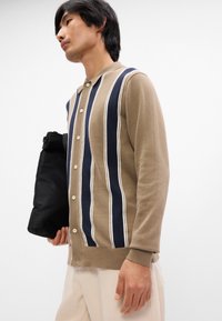 Beige kavaj med vertikala marinblåa och vita ränder, knappdesign framtill, ribbad krage och ärmslut, matchad med en svart väska.