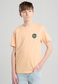 Persikka puuvillapaita, jossa on pyöreä kaula-aukko ja lyhyet hihat, ja jossa on vihreä ja musta brodeerattu "Löydä rauha" -grafiikka rinnassa.