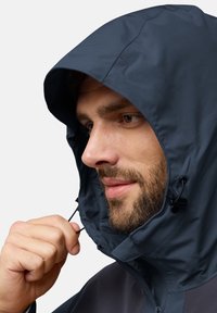 Veste imperméable bleu marine avec capuche, dotée de cordons réglables, d'une texture lisse et d'une fermeture éclair à l'avant. Vue rapprochée d'un modèle.