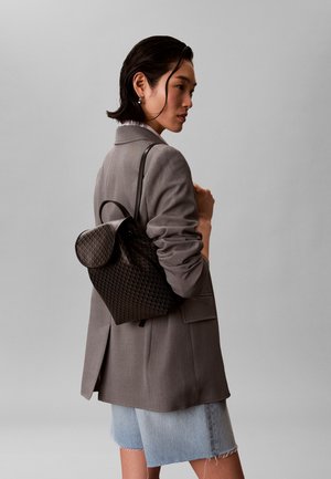 Ženska s kratkimi temnimi lasmi, oblečena v sivo jakno in jeans kratke hlače, nosi črno teksturirano nahrbtnik čez eno ramo na enakomerno sivi podlagi.