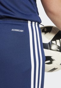 Marina sportshorts med vita vertikala ränder och "AEROREADY"-logotyp på midjebandet; håller en svart och vit fotboll.