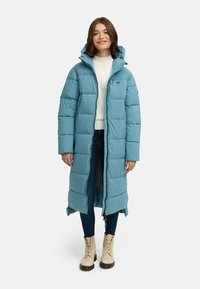 Lys blå lang dunjakke med hette, vertikal quiltet design, front glidelås, brukt over en hvit genser og mørke jeans, kombinert med beige støvler.