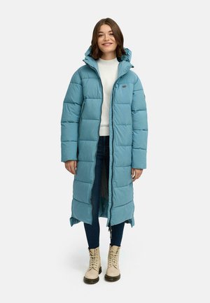 Lys blå lang dunjakke med hette, vertikal quiltet design, front glidelås, brukt over en hvit genser og mørke jeans, kombinert med beige støvler.