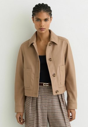 Femeie cu părul împletit poartă o jachetă bej cu nasturi, un top negru și pantaloni în carouri cu talie înaltă și curea, pe un fundal alb.