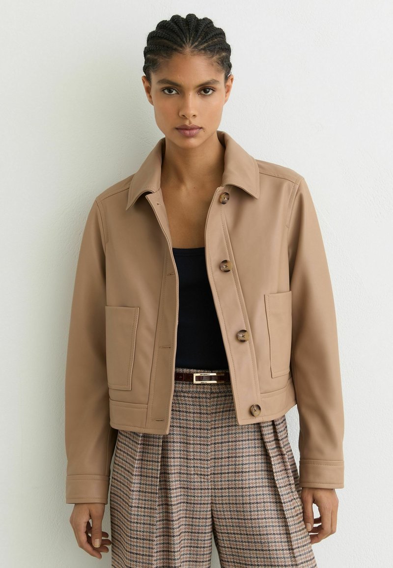 Žena s pletenými vlasmi má na sebe béžovú zapínaciu bundu, čierny vršok a kárované nohavice s vysokým pásom a opaskom na bielom pozadí.