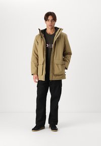 Khaki waterdichte jas met een capuchon, ritsluiting, twee voorkeuzes, zwarte voering. Gecombineerd met zwarte cargobroek en donkere schoenen.